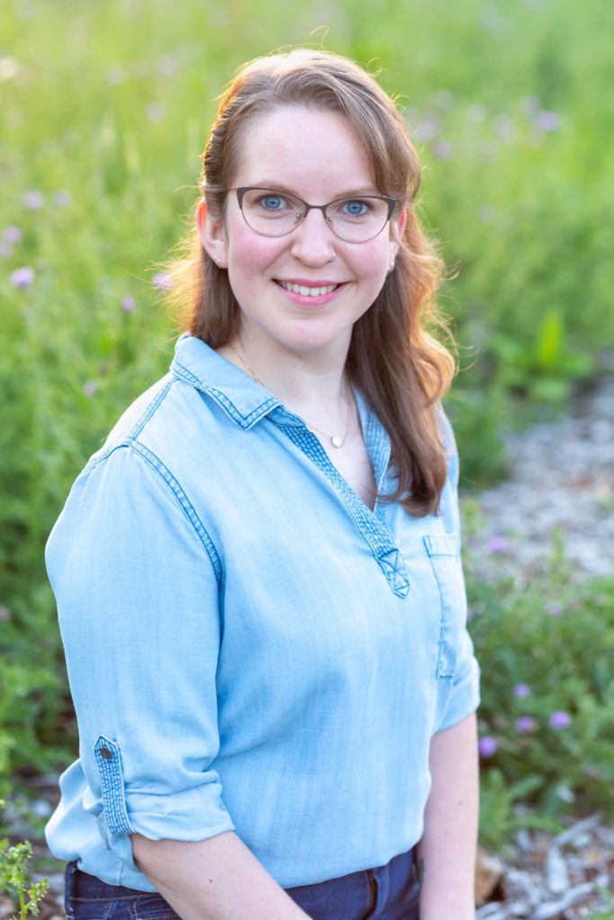 A young white women with light brown hair and glasses wearing a denim shirt sitting in the grass with the sun behind her.