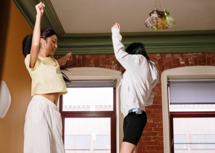 Two teenage girls standing on a bed dancing with their arms high in the air